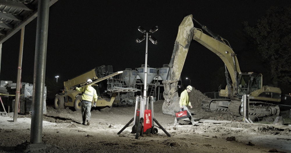 Iluminare de șantier pe timp de noapte cu turn de lumină Milwaukee alimentat cu baterie, într-un mediu de lucru industrial cu excavatoare.