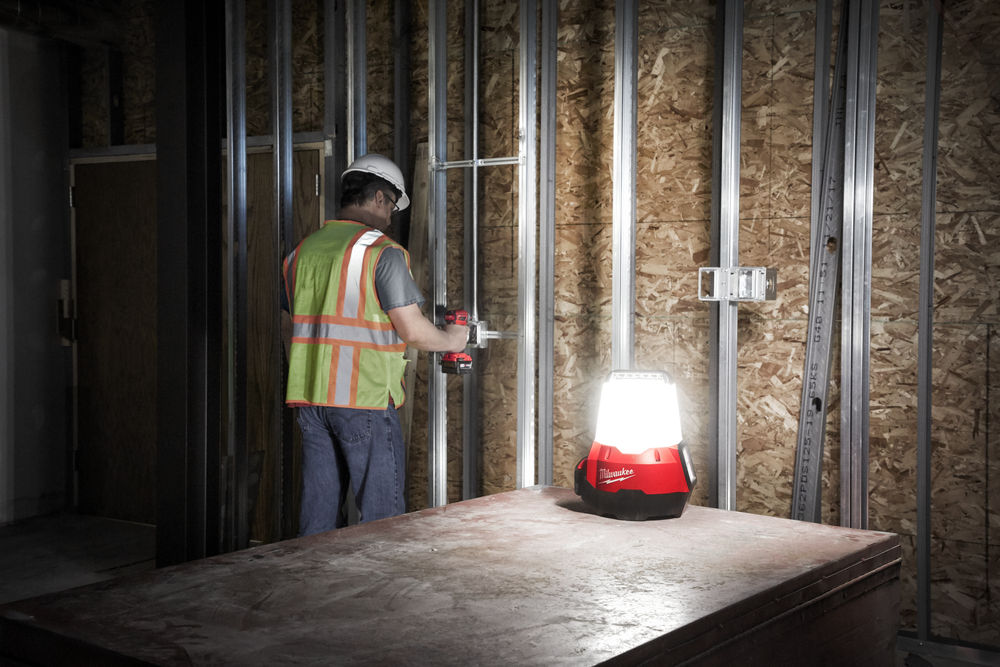 Lampă portabilă Milwaukee utilizată pentru iluminarea zonei de lucru, în timp ce un muncitor fixează profile metalice pe perete.