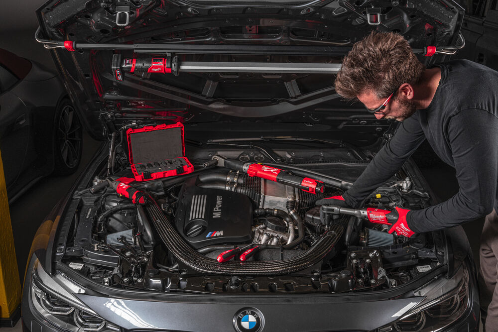 Un bărbat lucrează la motorul unei mașini BMW, folosind scule Milwaukee, concentrat pe detalii tehnice.