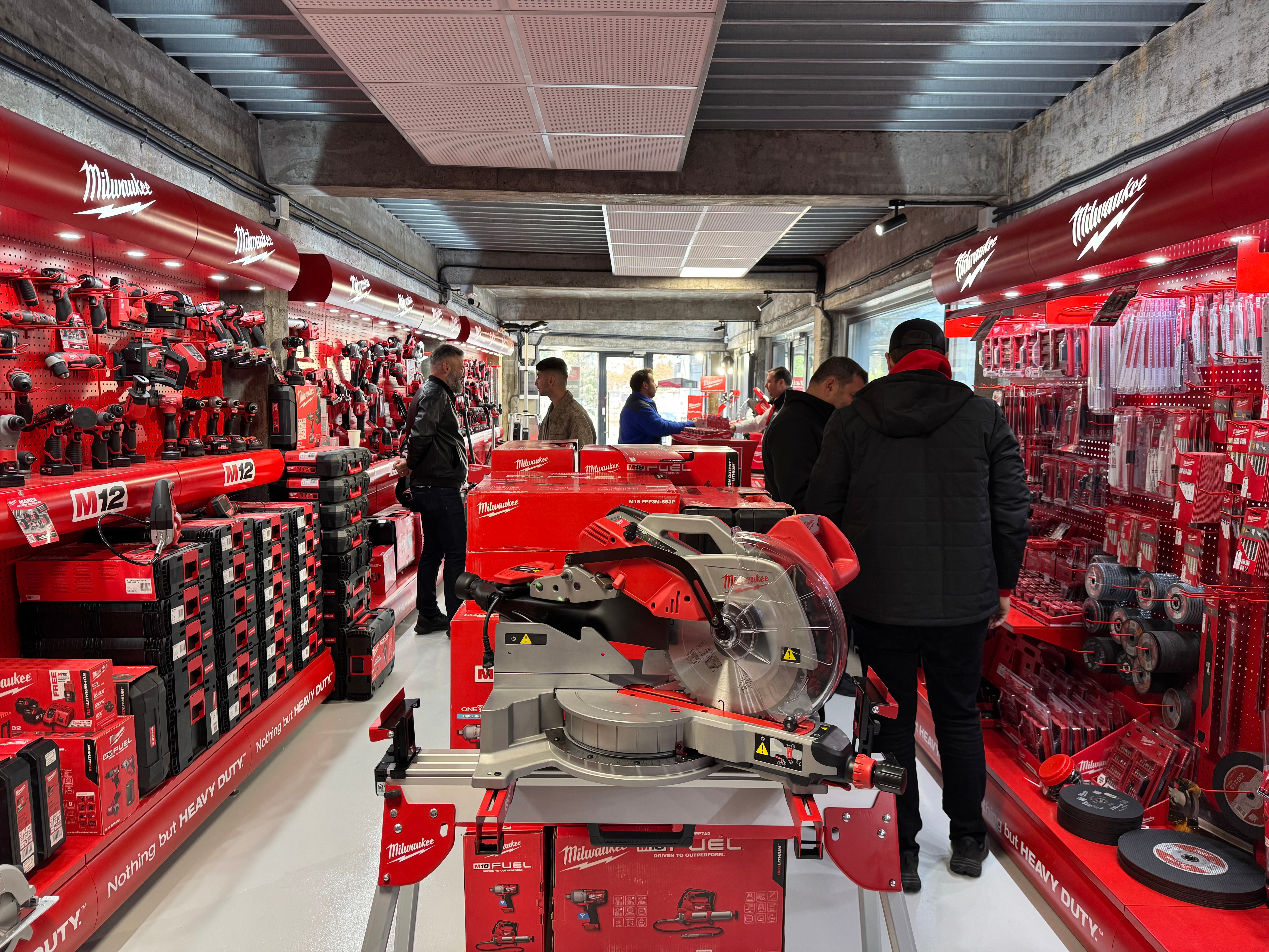 Showroom modern Iftode Universal dedicat sculelor Milwaukee, cu un perete întreg de unelte electrice și accesorii profesionale. Spațiul este organizat impecabil, evidențiind gama M12 și M18, cu standuri pline de bormașini, polizoare și fierăstraie circulare. Clienți și consultanți discută în mijlocul magazinului, într-un ambient industrial, dar curat și bine luminat.