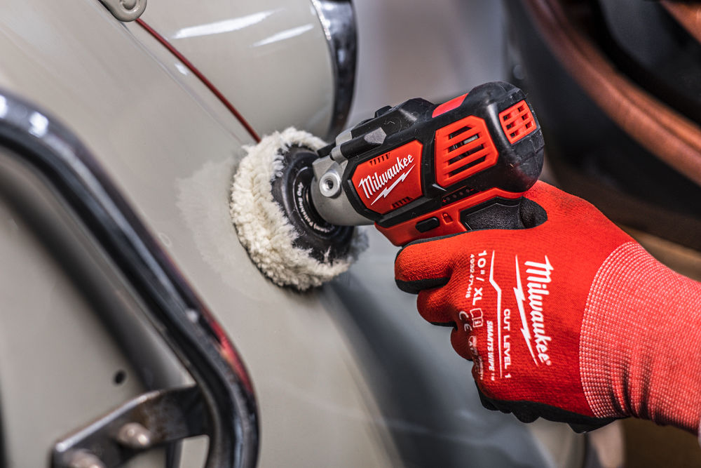 O persoană cu mănuși roșii Milwaukee folosește un șlefuitor fără fir cu disc textil pentru a lustrui caroseria unui automobil clasic.