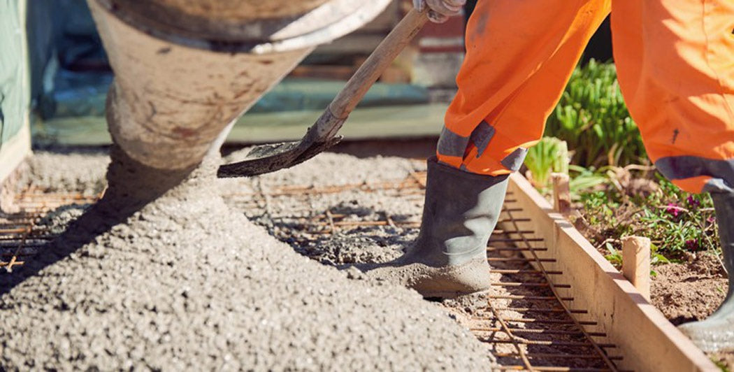 Turnare de beton proaspăt pe fundație, muncitor în echipament de protecție și cizme dirijează fluxul cu lopata.