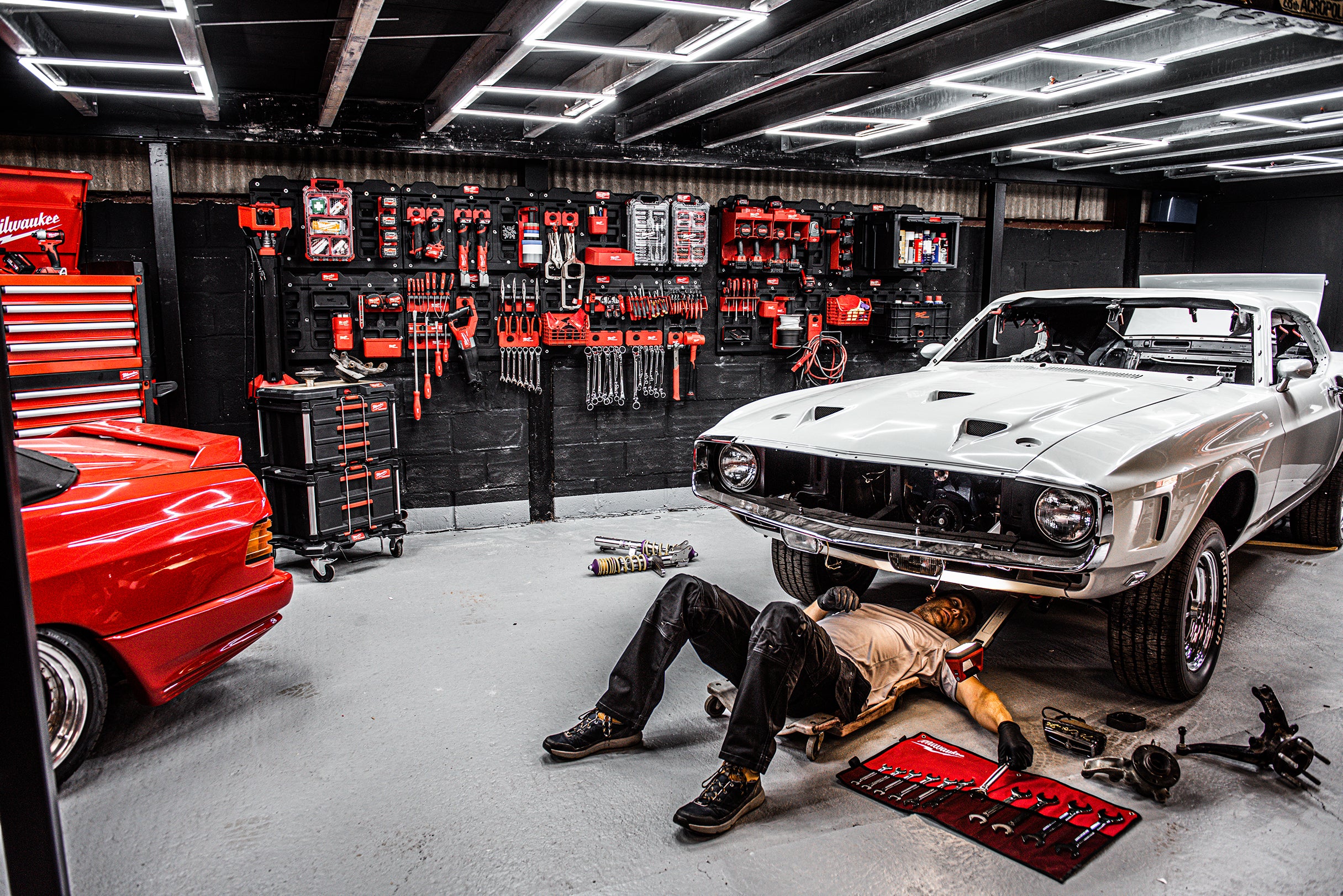 Un bărbat lucrează la o mașină într-un garaj, folosind scule Milwaukee pentru a efectua reparații