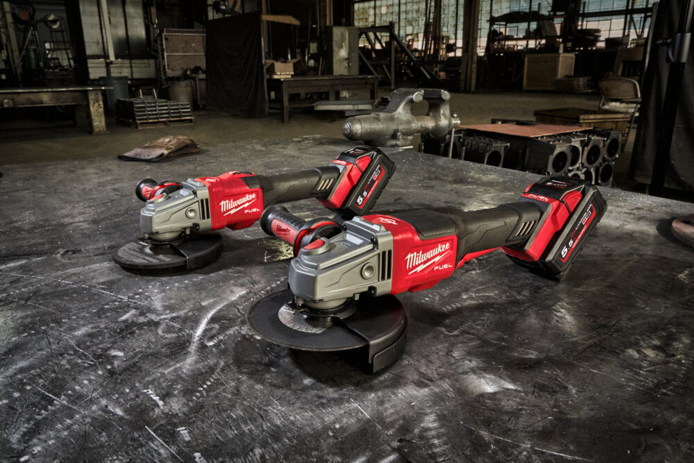 Două polizoare unghiulare Milwaukee M18 FUEL pe o masă metalică într-un atelier industrial, cu acumulatori REDLITHIUM 5.0 Ah.