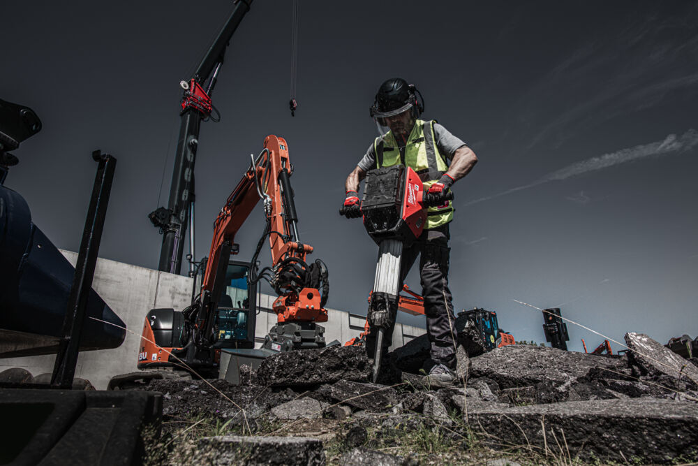 Muncitor sparge dale de asfalt folosind un ciocan demolator Milwaukee MX FUEL, pe un șantier industrial dotat cu utilaje grele.